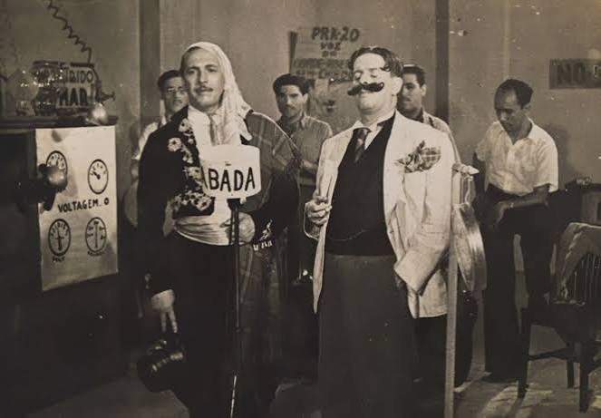 Fotografia em preto e branco de Castro Barbosa e Lauro Borges, radialistas, caracterizados em figurinos cômicos e diante de microfones, em um estúdio de rádio. Ao fundo, músicos e equipamentos de transmissão.